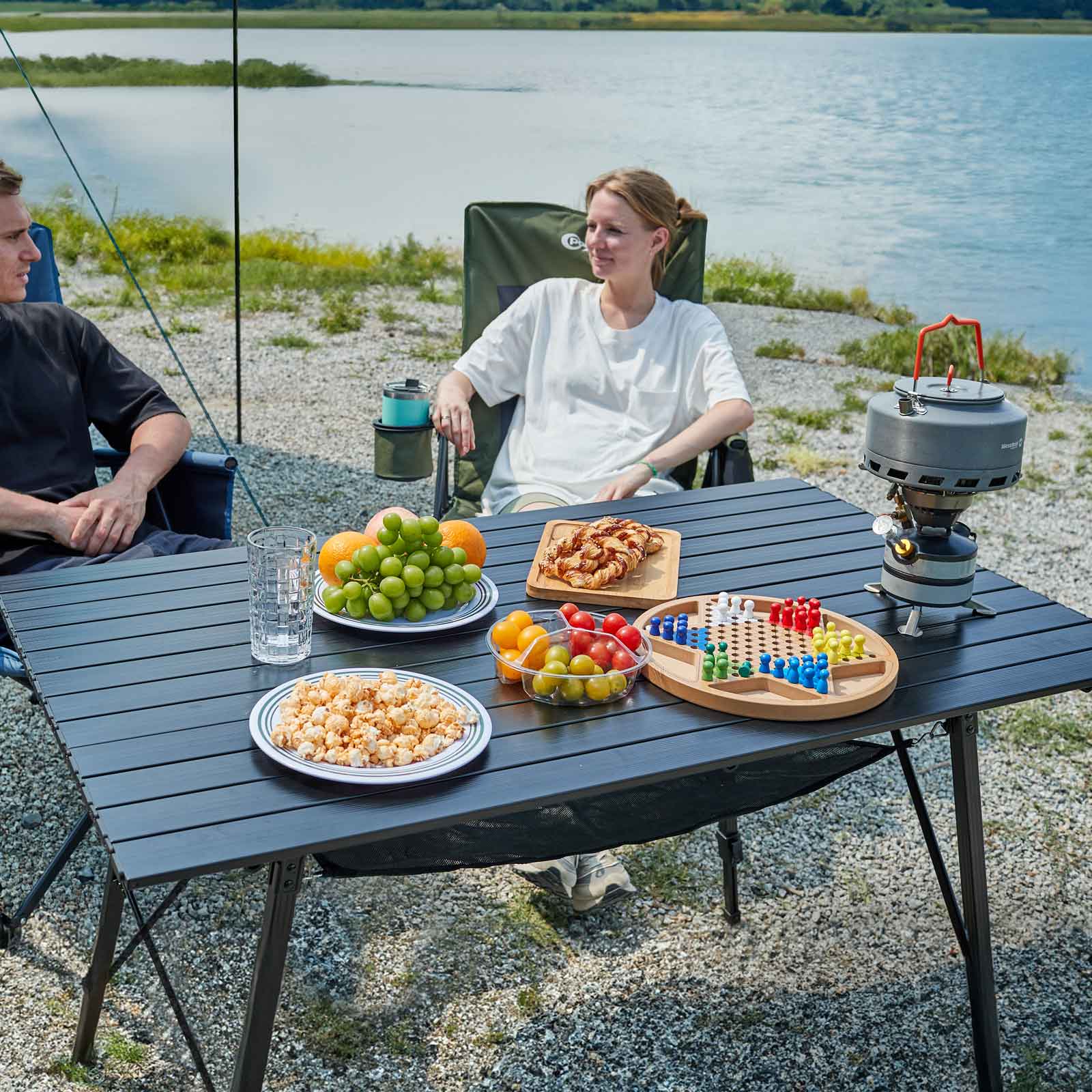 Two people sitting at a Portal Outdoors camping table by a lake, enjoying snacks and a board game.